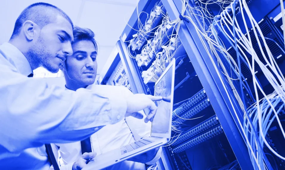 Customer working on networking technology while being assisted by another co-worker. Both of them are referencing a laptop for guidance.